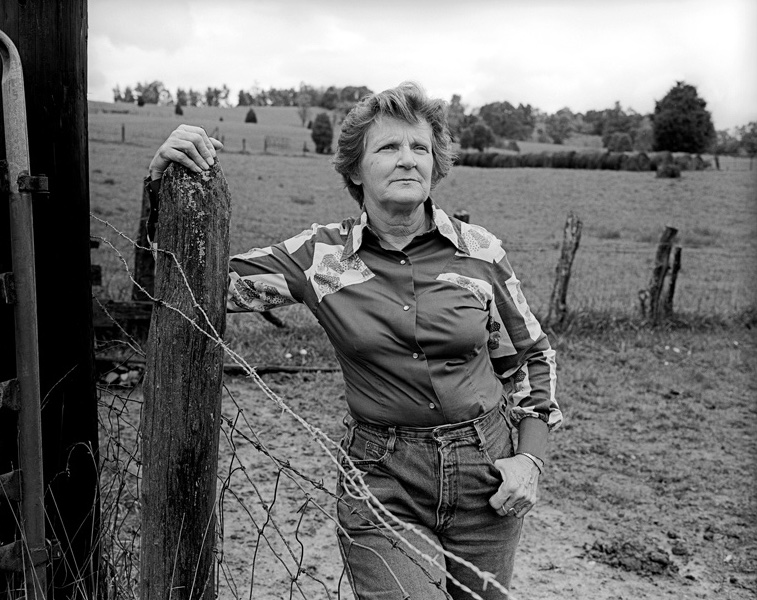Black-and-white photograph by Barbara Beirne. Mary Jack Hargins stands outside next to a wooden post and a metal wire fence. She's a middle-aged white woman with short hair, wearing a Western long-sleeve shirt and jeans. Her left hand rests on the wooden post, and her right thumb is hooked on her pocket.
