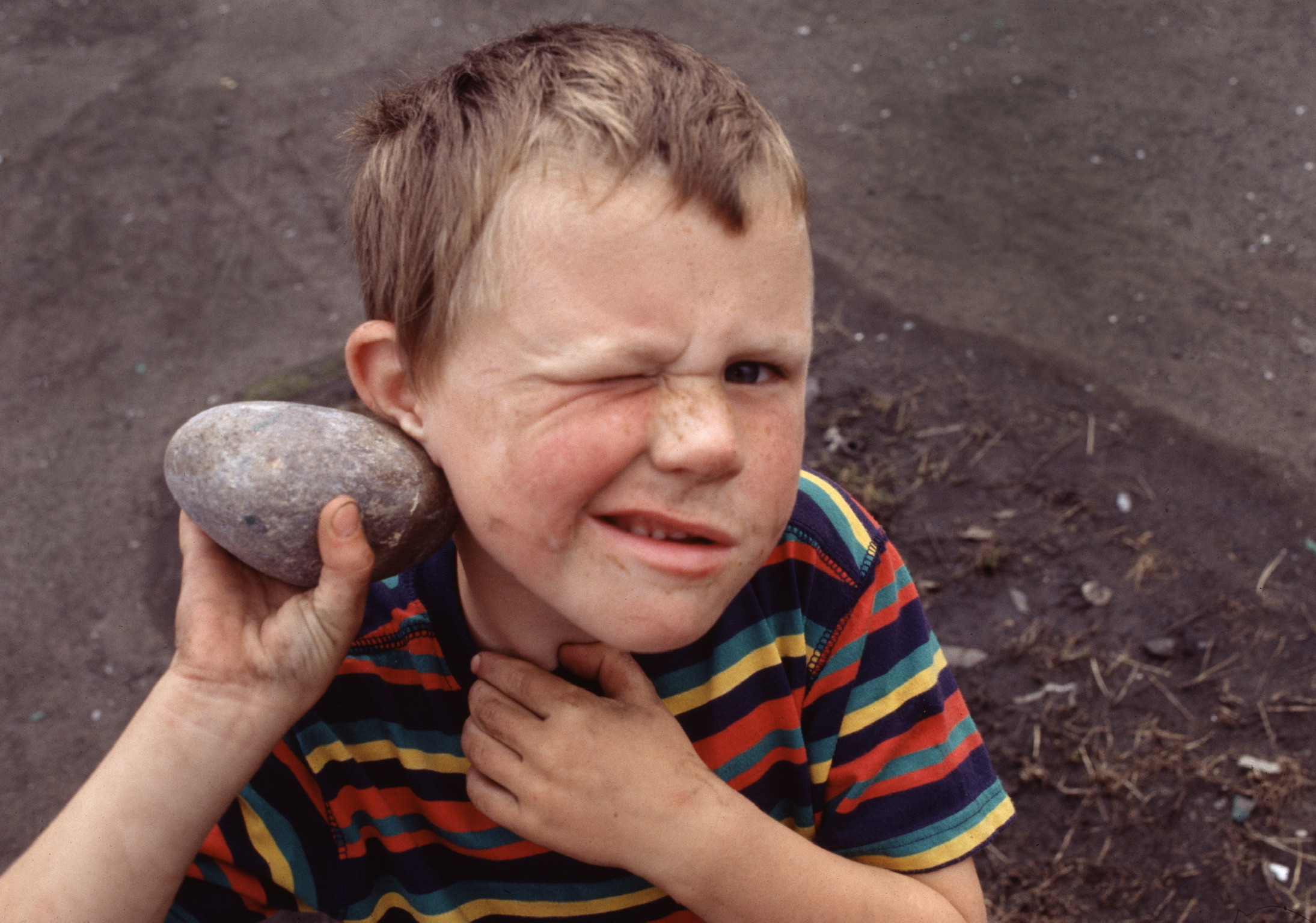 Color photograph by Barbara Beirne. High-angle shot looking down onto a young boy holding a rock in his left hand, close to his ear. His right eye is closed, and his mouth curled up to the side. He wears a striped T-shirt.