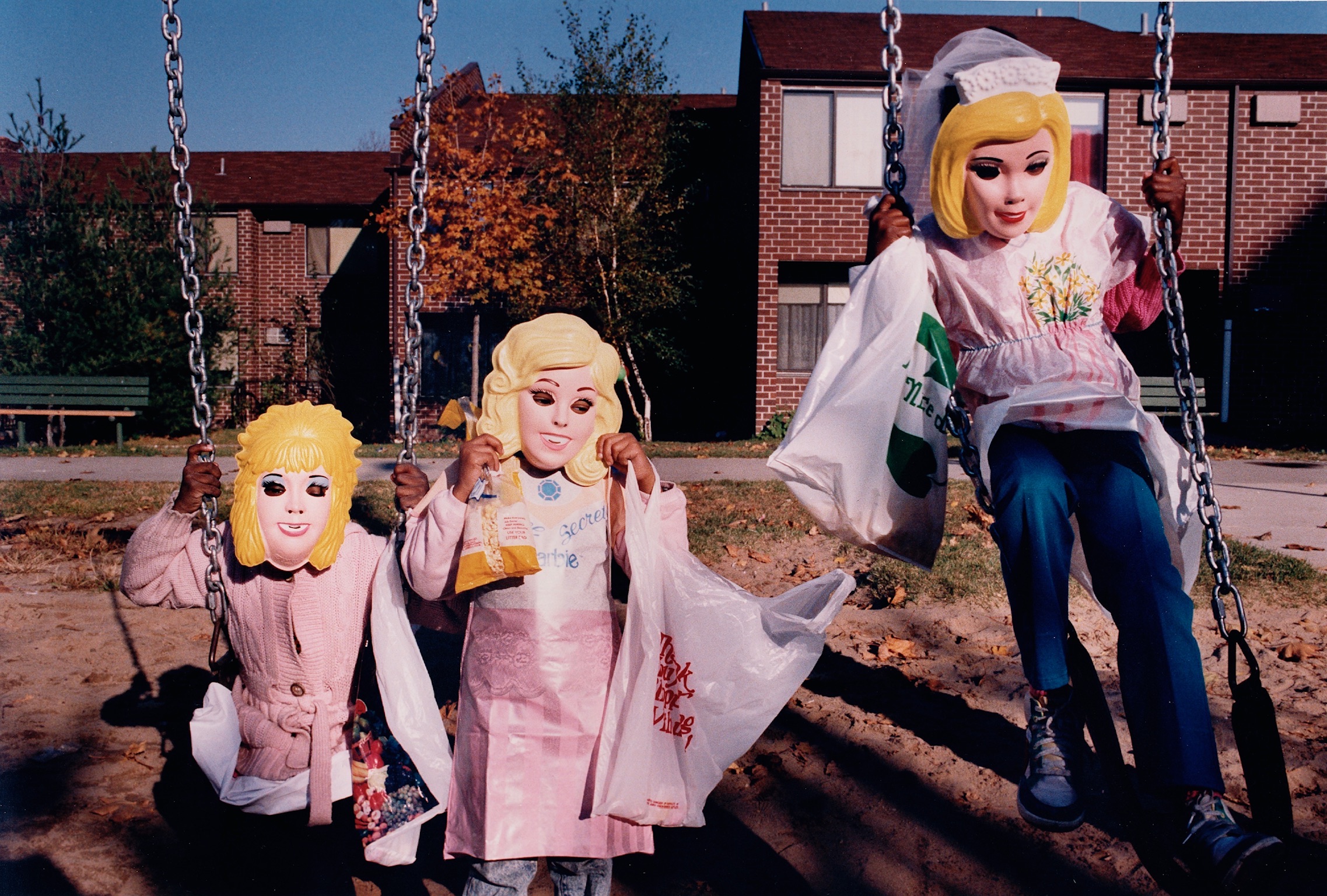Color photograph by Barbara Beirne. Three young black girls in a playground wearing pink costumes and plastic masks of blonde white women.
                        