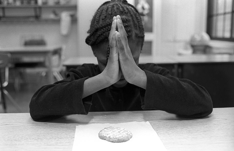 Black-and-white photograph by Barbara Beirne. A black child sits at the table before a cookie on a paper napkin, head down, holding her hands in prayer.
                    